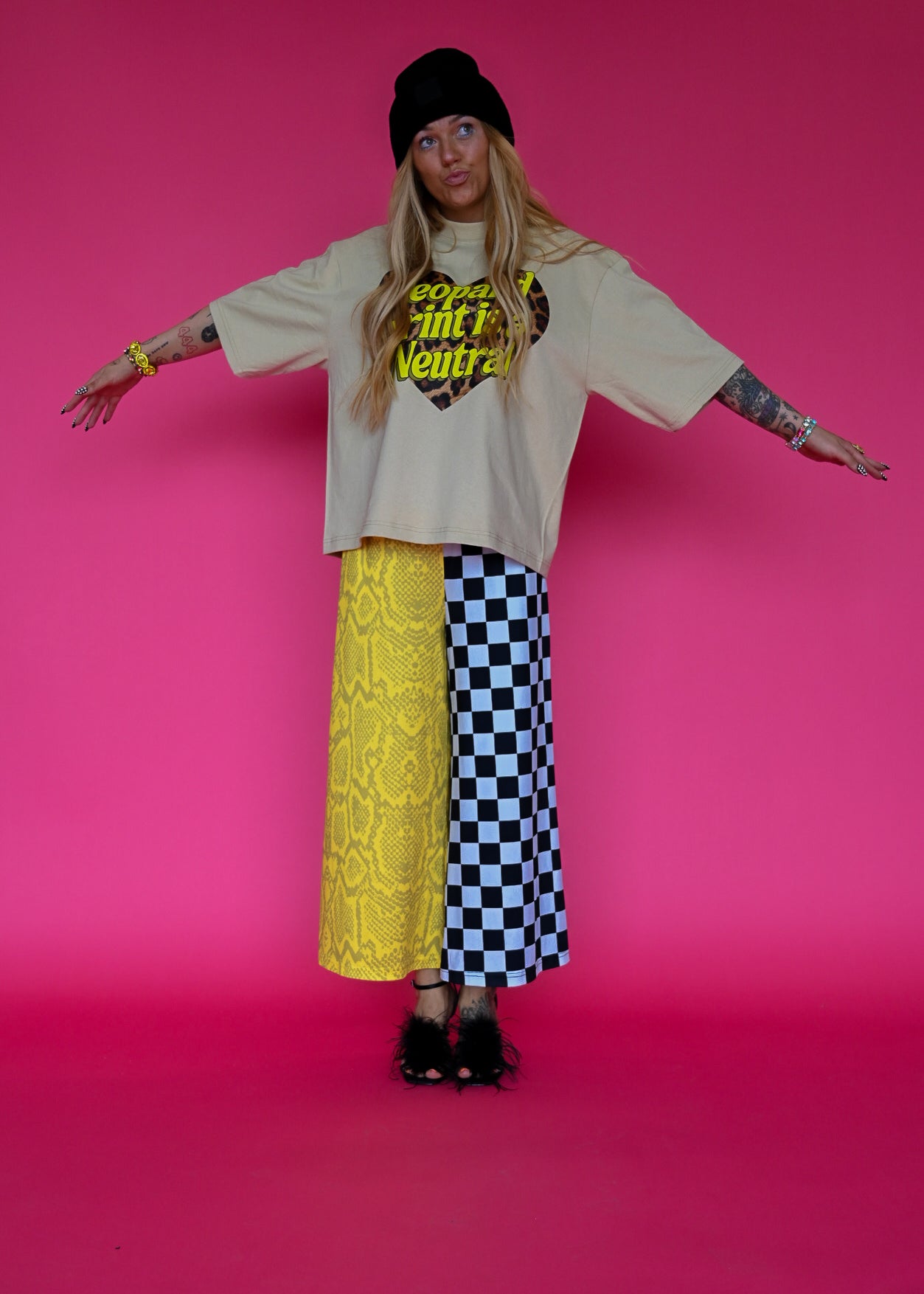 A model stands against a solid pink studio background wearing a beige graphic t-shirt, a yellow lace skirt on one side and a black and white checkered pants on the other, a black beanie, and black heels
