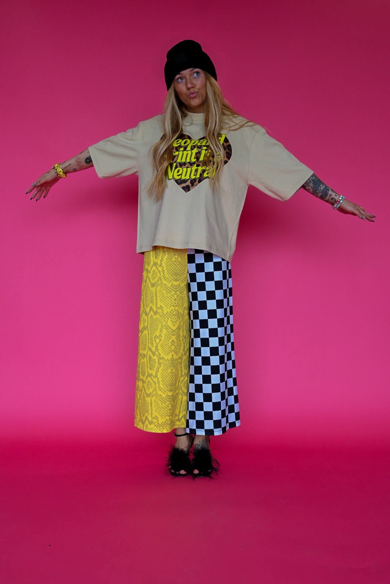 A model stands against a solid pink studio background wearing a beige graphic t-shirt, a yellow lace skirt on one side and a black and white checkered pants on the other, a black beanie, and black heels