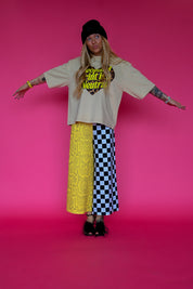 A model stands against a solid pink studio background wearing a beige graphic t-shirt, a yellow lace skirt on one side and a black and white checkered pants on the other, a black beanie, and black heels