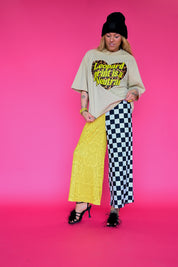 A model stands against a solid pink studio background wearing a beige graphic t-shirt with a leopard print heart, a bright yellow lace skirt on one leg, and black-and-white checkered pants on the other leg, paired with a black beanie and black heels