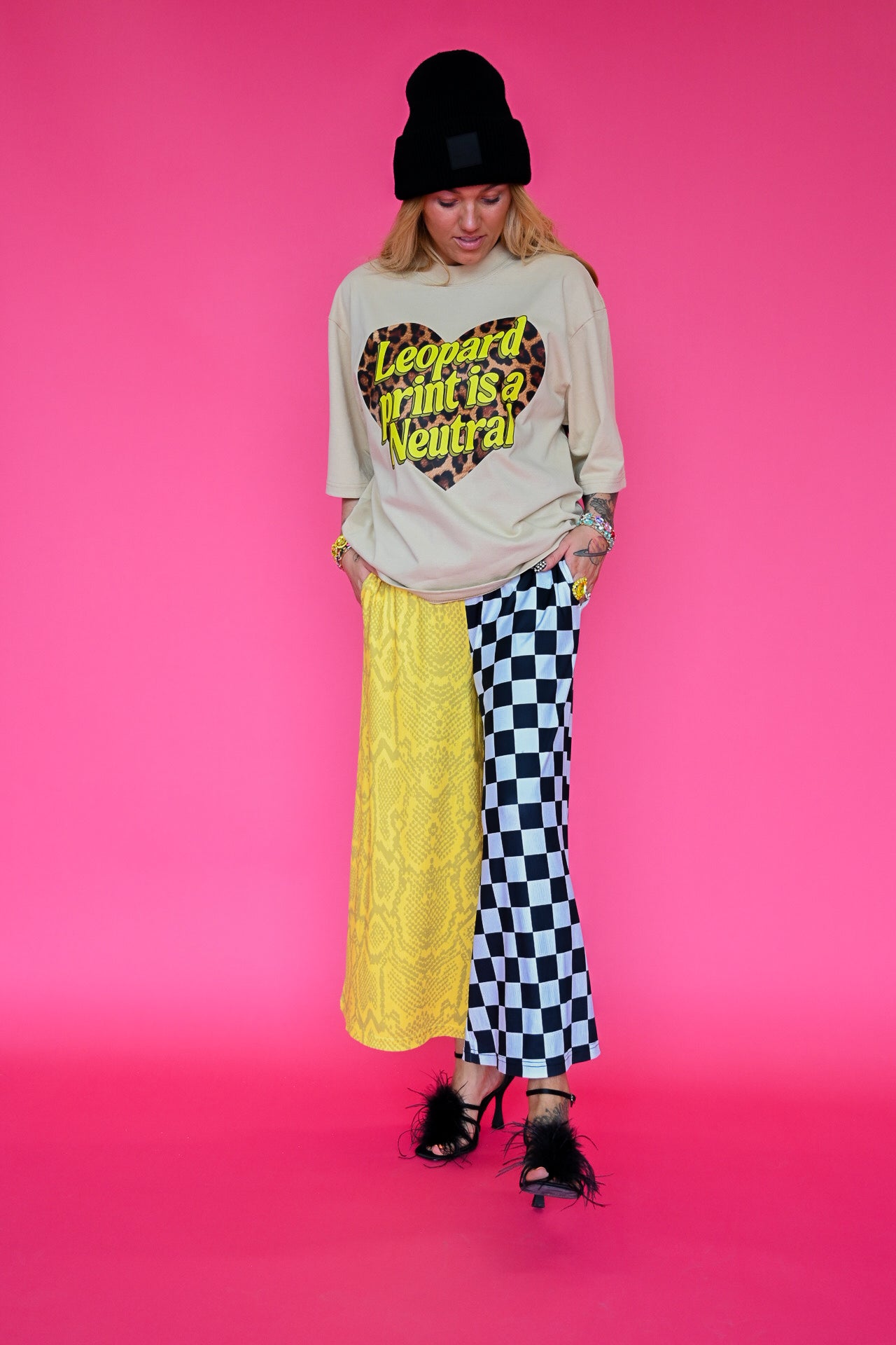 A model wearing a beige graphic tee that says 'leopard print is a neutral' and half and half pants that are yellow snakeskin, and black & white checkered patterned, on a pink background