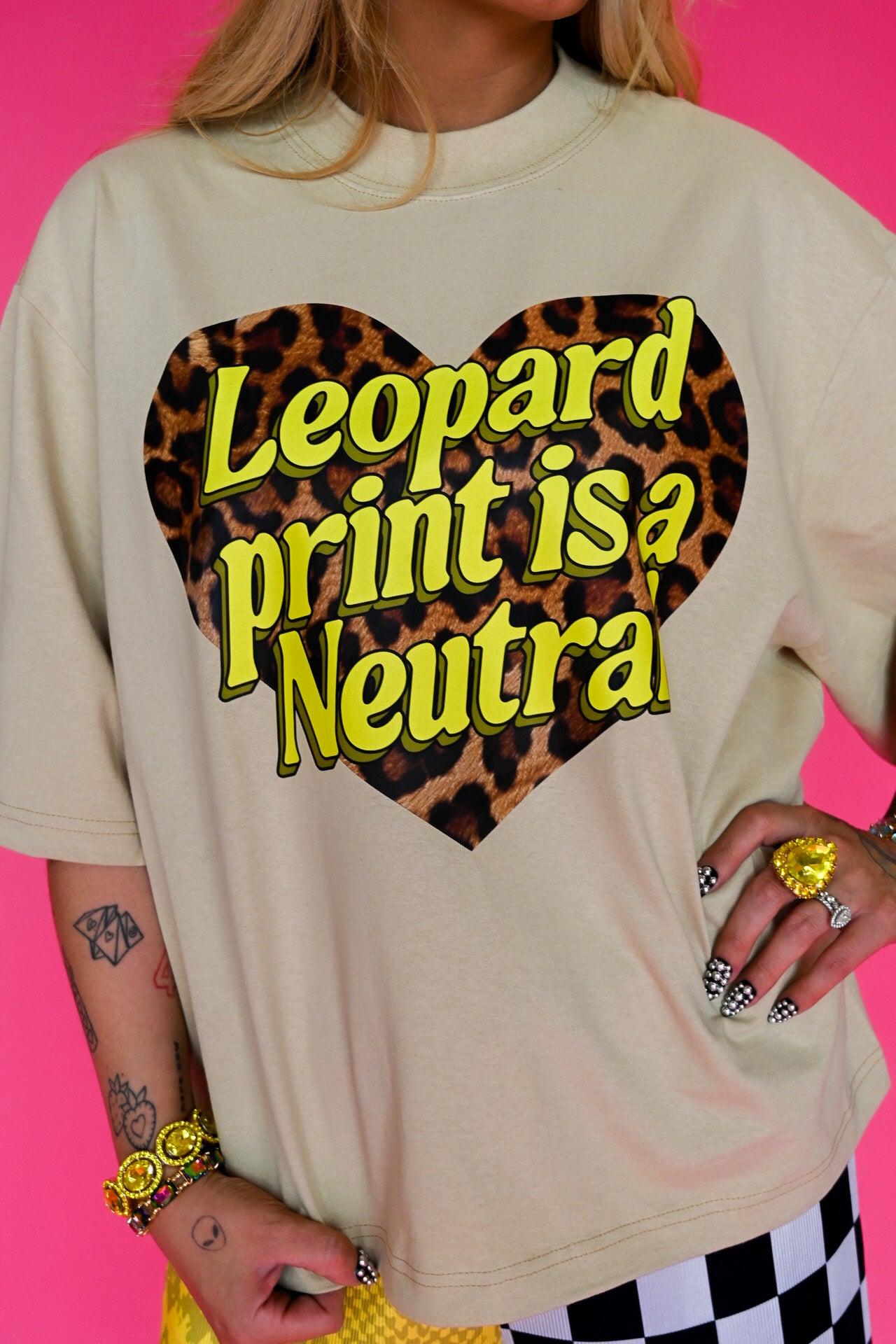 A person wearing an oversized cream-colored t-shirt with a large leopard-print heart graphic and the text 'Leopard print is a Neutra' in yellow. The background is a solid bright pink