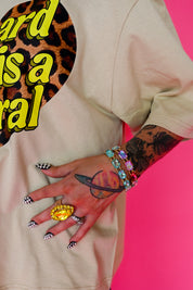 A close-up lifestyle shot of a person wearing a beige oversized t-shirt with a graphic print, featuring a leopard print circle and bold yellow text. The person has a Saturn tattoo on their hand, colorful gemstone bracelets, and a large gold ring, posed against a vibrant hot pink background