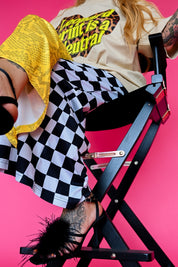 A model sitting in a chair wearing a beige graphic tee that says 'leopard print is a neutral' and half and half pants that are yellow snakeskin, and black & white checkered patterned, on a pink background, focus on the pant leg and black fuzzy heals