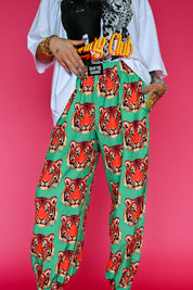A person wearing green harem-style pants with a repeating red and orange tiger face print, styled with a white graphic T-shirt and gold jewelry against a solid pink background