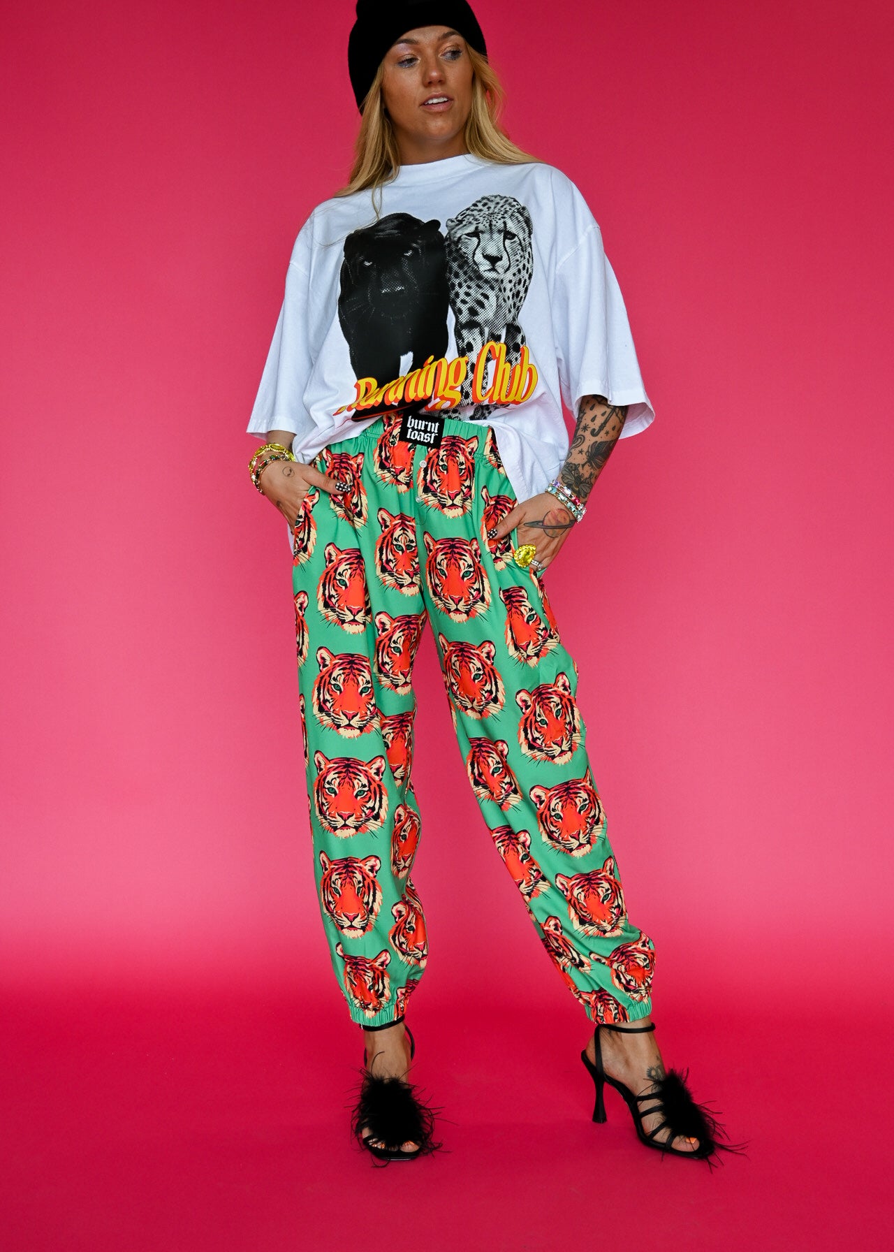 A model stands against a solid pink studio background wearing a white graphic t-shirt with a black leopard print and yellow 'Running Club' text, paired with green harem-style pants featuring a red tiger print, a black beanie, and black strappy heels