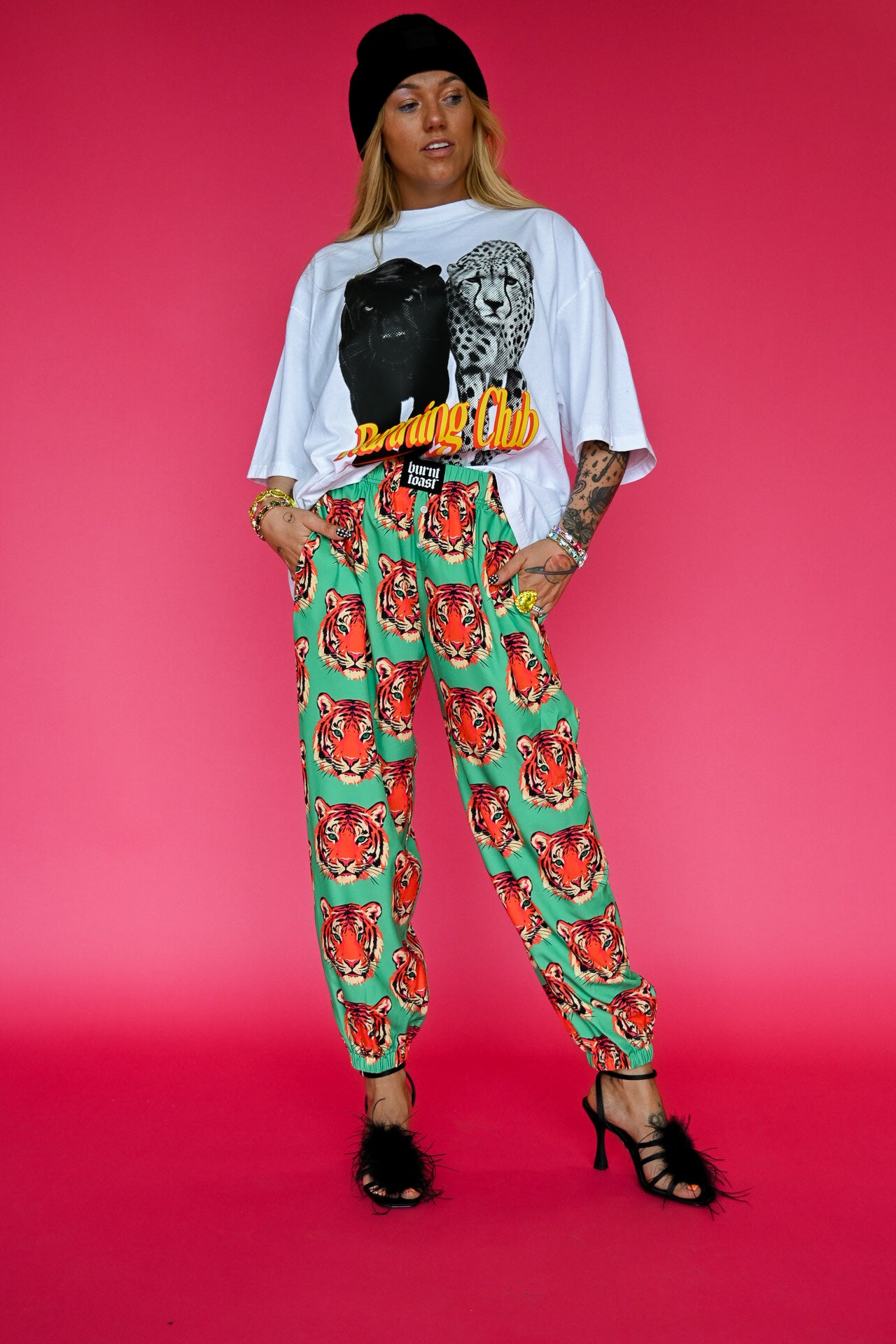 A model stands against a solid pink studio background wearing a white graphic t-shirt with a black leopard print and yellow 'Running Club' text, paired with green harem-style pants featuring a red tiger print, a black beanie, and black strappy heels