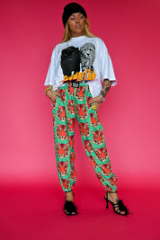 A model stands against a solid pink studio background wearing a white graphic t-shirt with a black leopard print and yellow 'Running Club' text, paired with green harem-style pants featuring a red tiger print, a black beanie, and black strappy heels