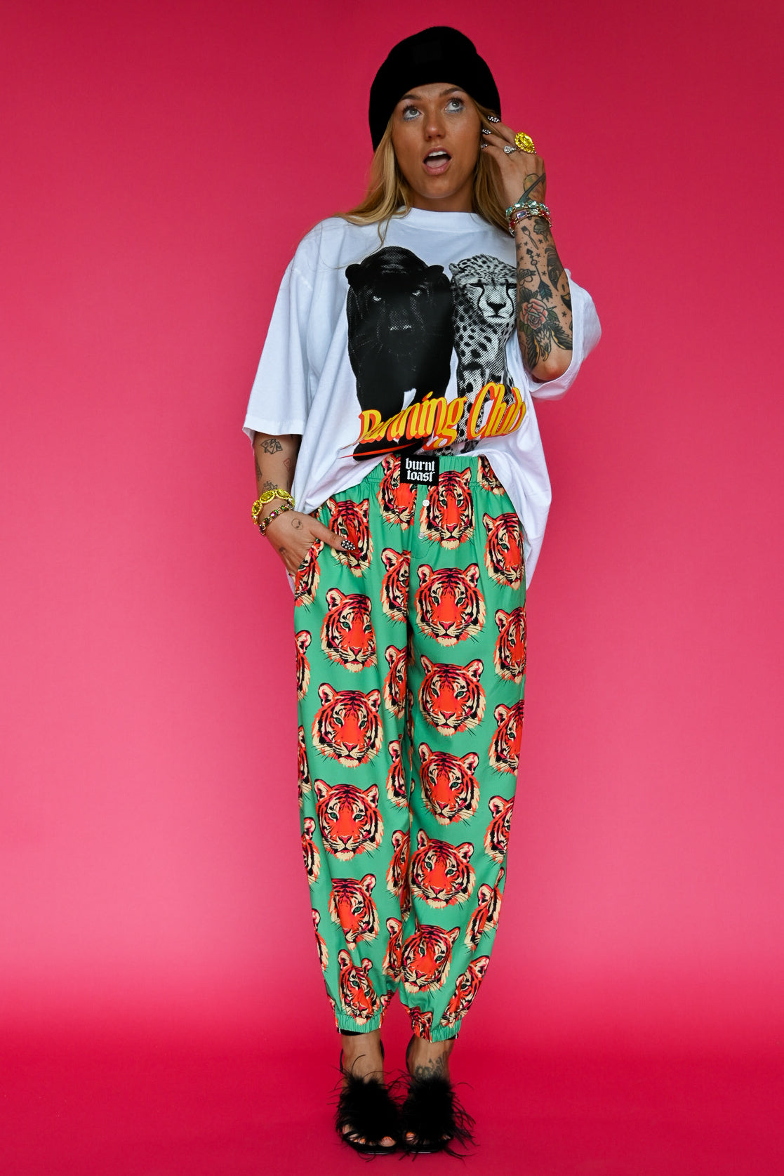 A model stands against a vibrant pink studio background wearing a white graphic t-shirt, a black beanie, a black crossbody bag, and green pants with a red and orange tiger print