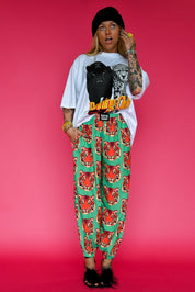 A model stands against a vibrant pink studio background wearing a white graphic t-shirt, a black beanie, a black crossbody bag, and green pants with a red and orange tiger print