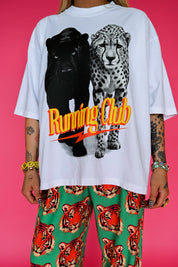A person wearing an oversized white graphic T-shirt with a black panther and spotted big cat print and the words 'Running Club' in orange/yellow, paired with green pants featuring a tiger face pattern, against a solid pink background