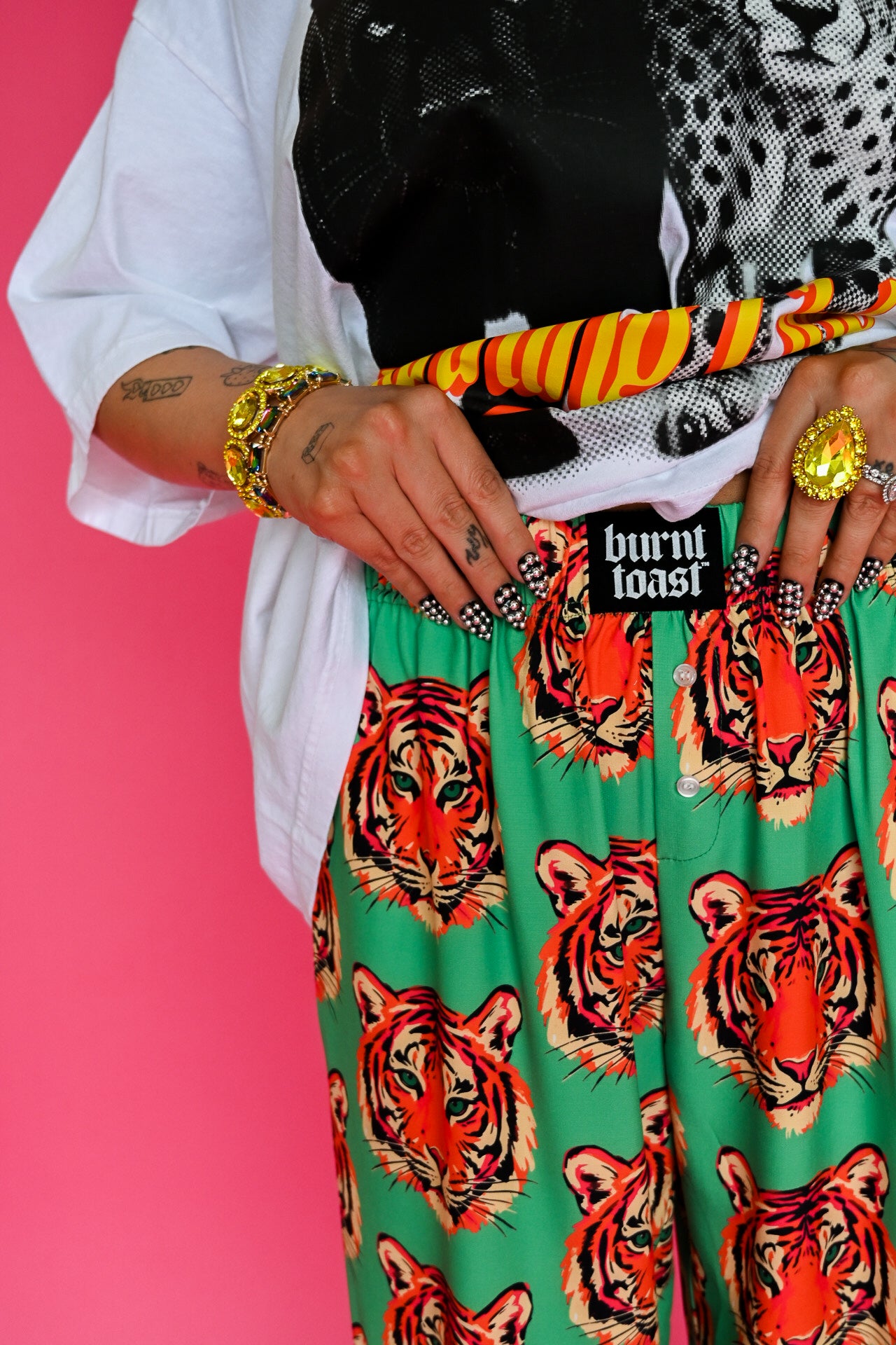 A close-up of a person wearing bright green trousers with a bold orange and black tiger print, styled with a white shirt, black top, patterned scarf, and gold jewelry against a solid pink background