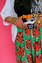 A close-up of a person wearing bright green trousers with a bold orange and black tiger print, styled with a white shirt, black top, patterned scarf, and gold jewelry against a solid pink background
