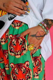 A close-up of a person wearing a bright green skirt with a bold red, orange, and black tiger print, styled with a white shirt and multiple colorful statement rings and bracelets