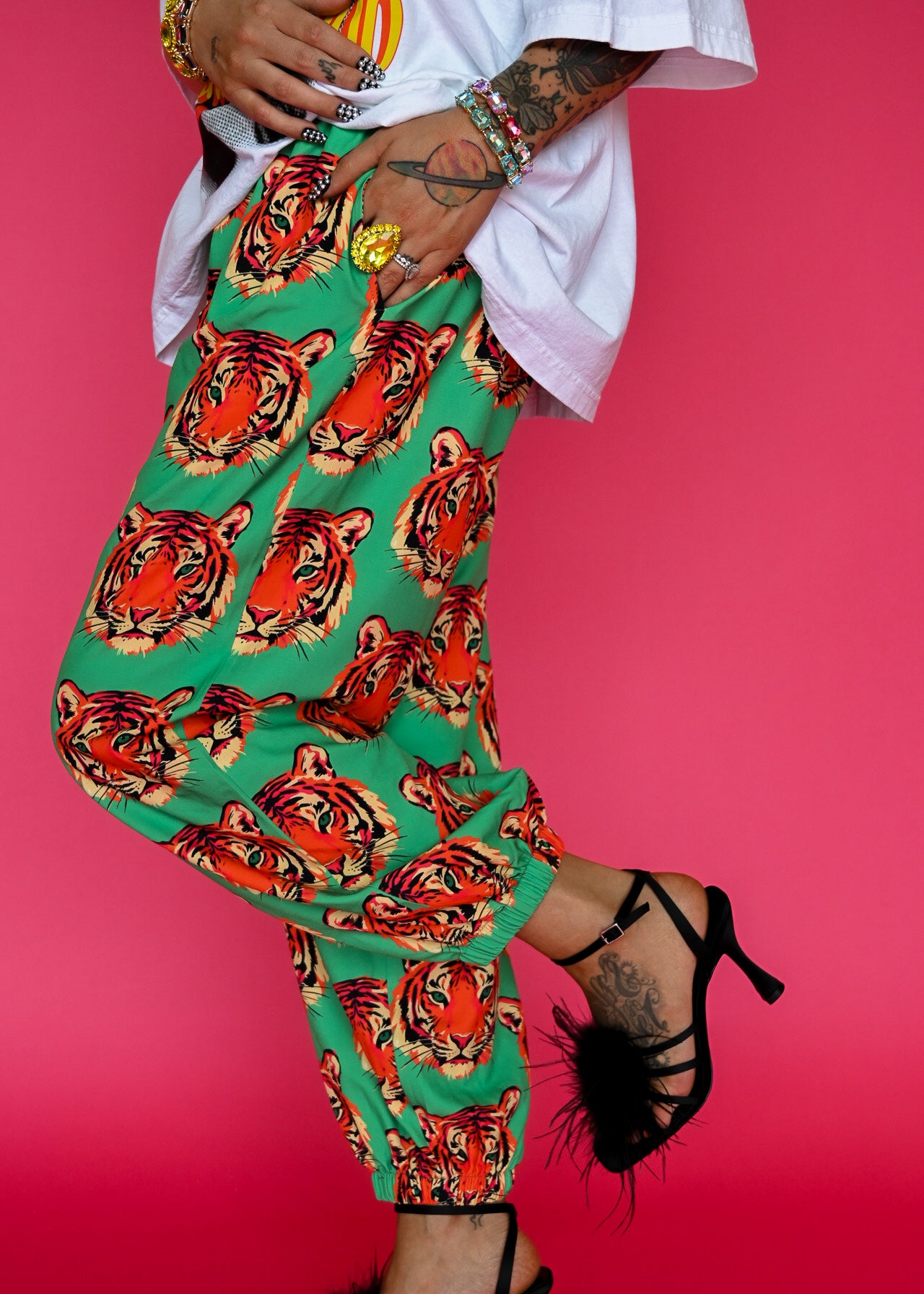 A person wearing bright green harem-style pants covered in a bold orange, red, black, and cream tiger print, paired with a white top and black high-heeled sandals, posed against a solid pink background