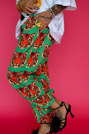 A person wearing green pants with a bold red and orange tiger face print, styled with a white shirt, gold jewelry, and black high-heeled sandals against a solid pink background