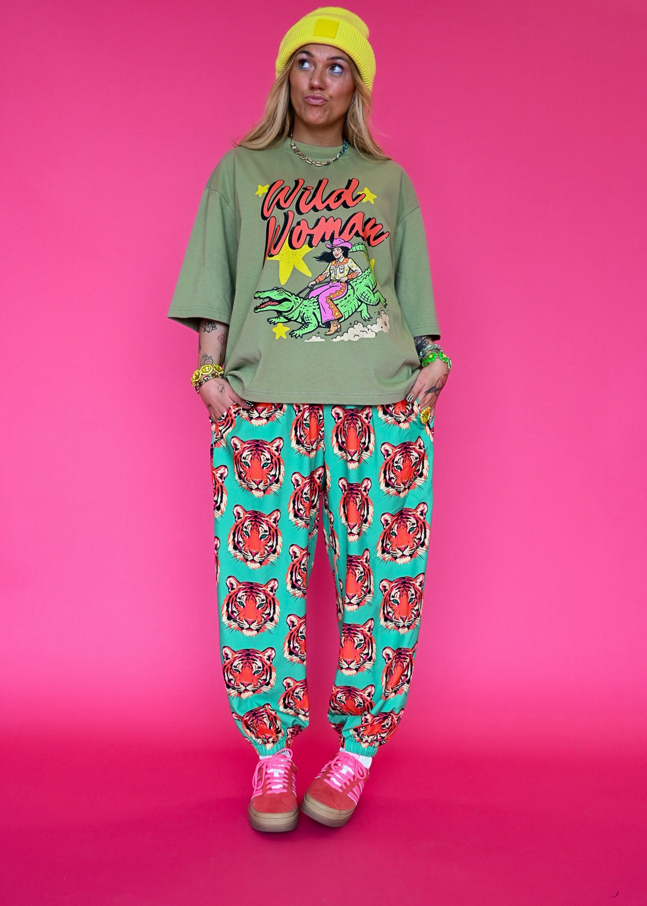 A model stands against a solid pink studio background wearing a green graphic t-shirt with 'Wild Woman' text, teal pants with a red tiger print, a yellow beanie, and pink sneakers