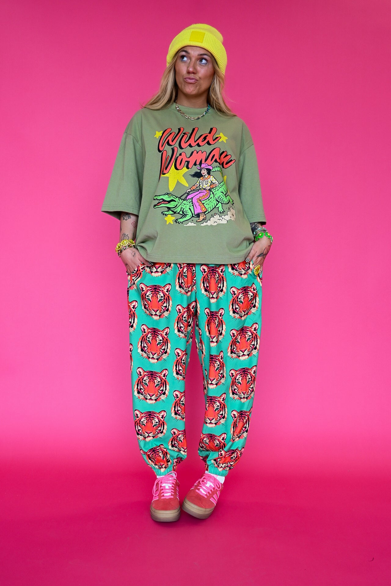 A model stands against a solid pink studio background wearing a green graphic t-shirt with 'Wild Woman' text, teal pants with a red tiger print, a yellow beanie, and pink sneakers