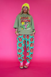 A model stands against a solid pink studio background wearing a green graphic t-shirt with 'Wild Woman' text, teal pants with a red tiger print, a yellow beanie, and pink sneakers