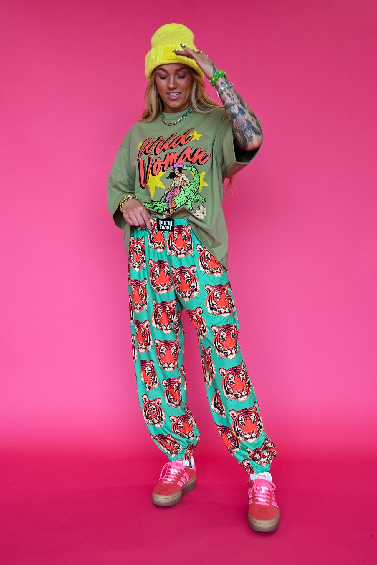 A model posing in a vibrant studio setting against a solid pink background, wearing a green graphic t-shirt, teal and red patterned sweatpants, a bright yellow beanie, and pink sneakers
