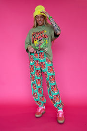 A model posing in a vibrant studio setting against a solid pink background, wearing a green graphic t-shirt, teal and red patterned sweatpants, a bright yellow beanie, and pink sneakers