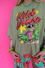 A person wearing an oversized sage green graphic T-shirt with a colorful 'Wild Woman' illustration on the front, paired with bright patterned pants, posed against a solid hot pink background