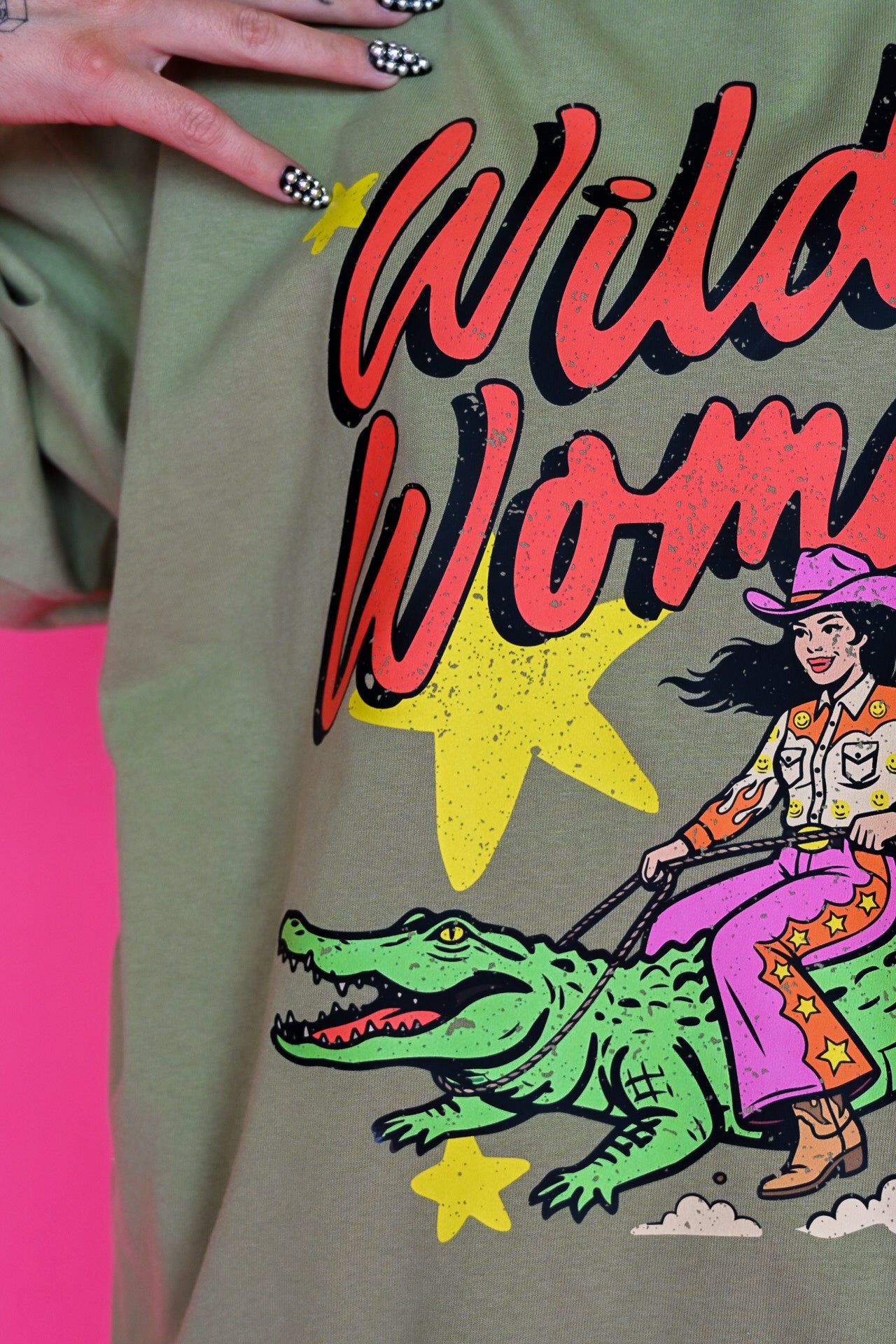A close-up of a person wearing an olive green graphic t-shirt. The shirt features a retro-style western print with the text 'Wild Woman' in red and black, yellow stars, a green alligator, and a cowboy riding it