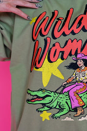 A close-up of a person wearing an olive green graphic t-shirt. The shirt features a retro-style western print with the text 'Wild Woman' in red and black, yellow stars, a green alligator, and a cowboy riding it