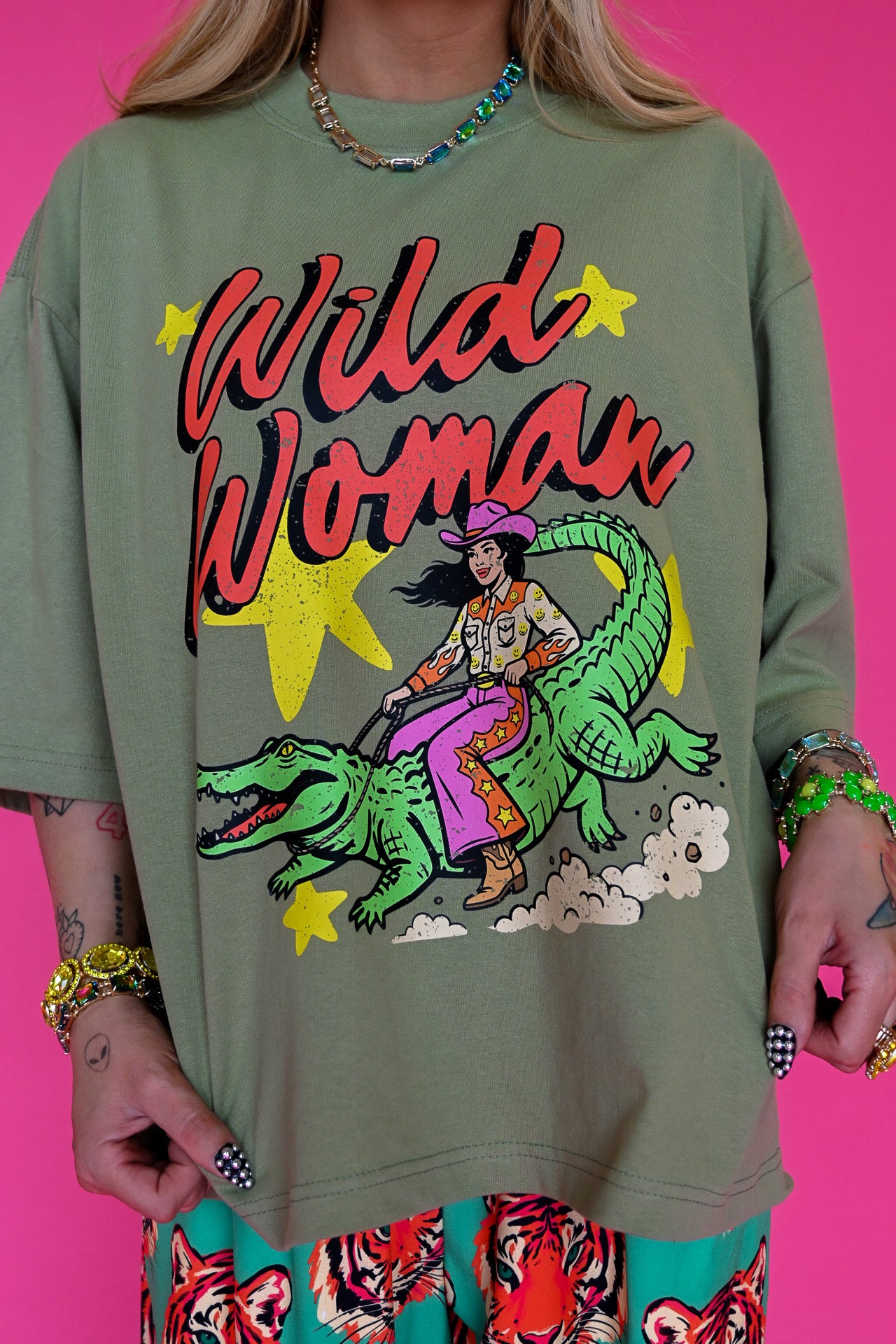 An oversized olive green graphic t-shirt featuring a retro-style illustration of a woman riding a green alligator with the text 'Wild Woman' in red and yellow. The model is wearing colorful patterned tiger-print pants against a solid bright pink background