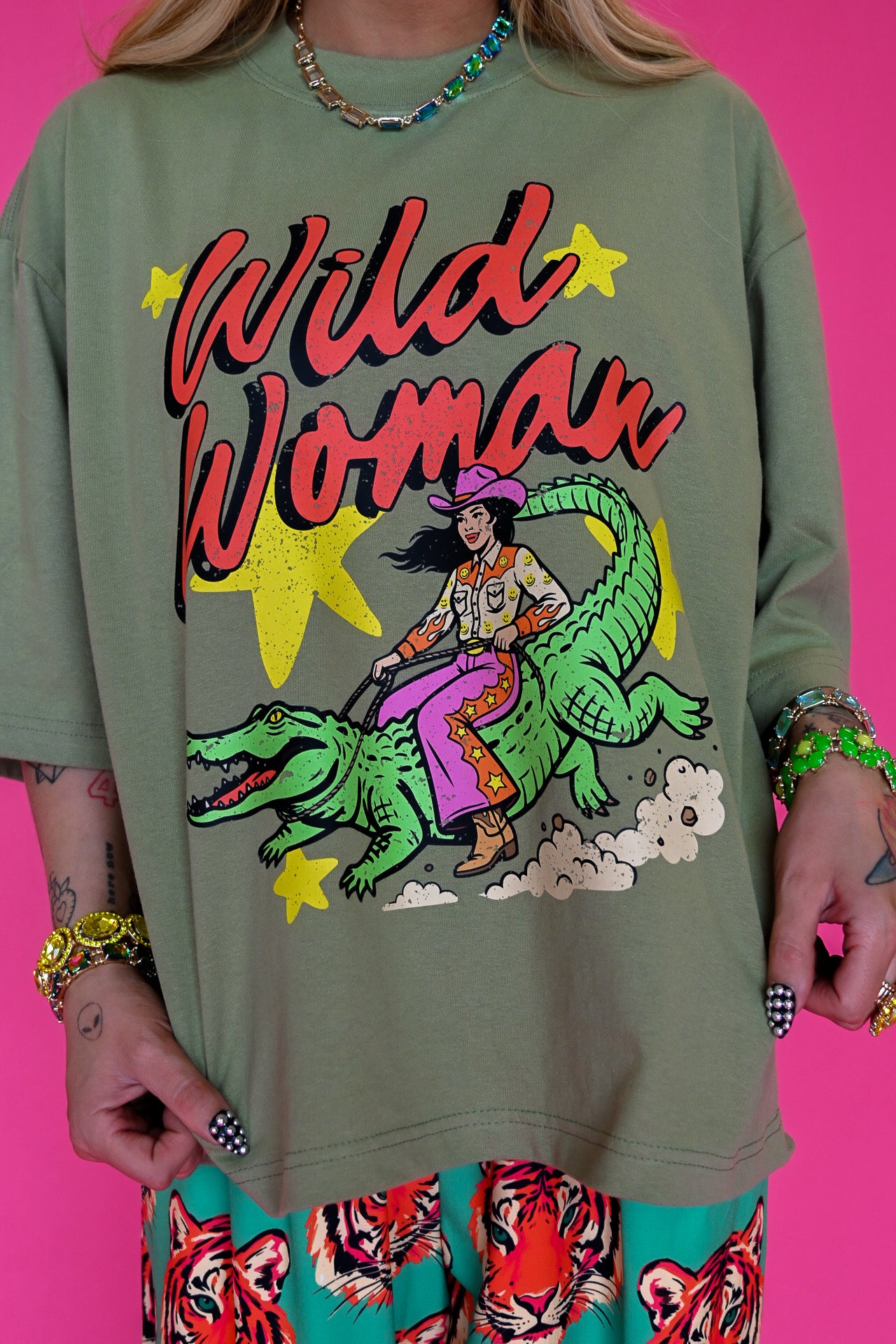 An oversized olive green graphic t-shirt featuring a colorful illustration of a woman riding a green alligator with the text 'Wild Woman' in red and yellow. The model is also wearing tiger-print pants and colorful jewelry, set against a solid bright pink background