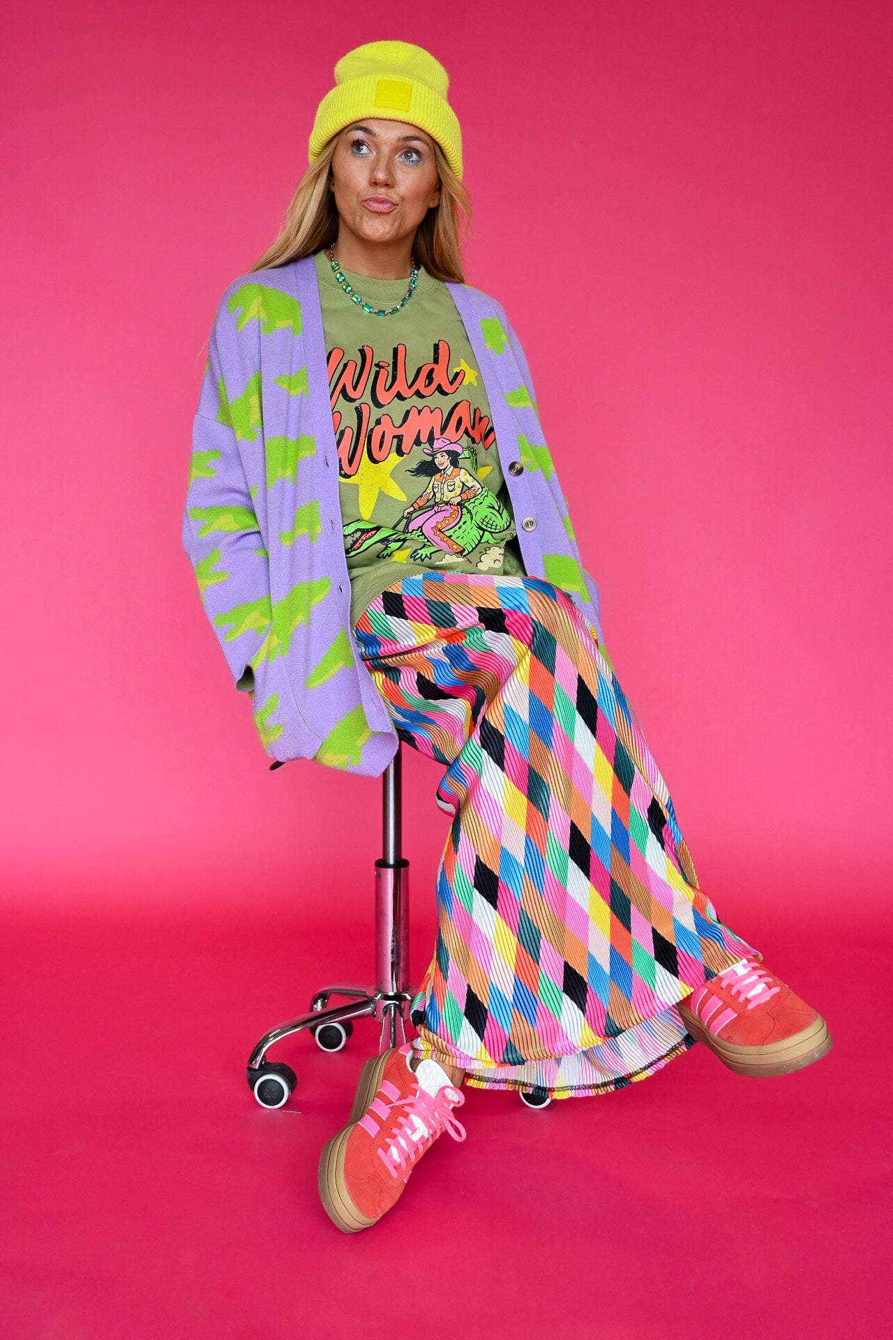 A model sits on a chrome rolling stool against a vibrant pink studio background, wearing a colorful retro-style outfit with a pastel tie-dye cardigan, a pink and yellow diamond-patterned skirt, a graphic t-shirt, and pink sneakers