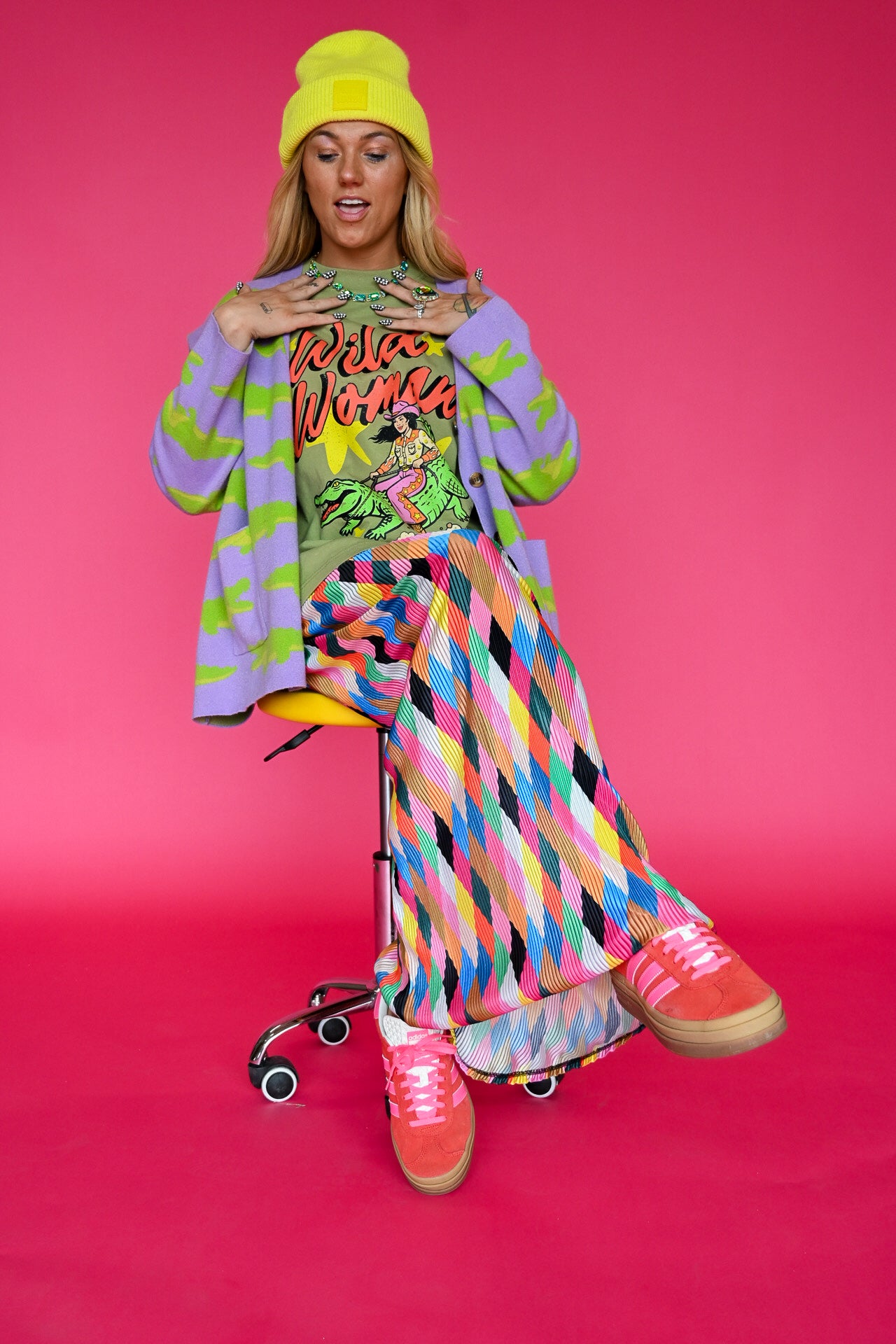A woman posing on a yellow rolling stool against a solid pink background, wearing a vibrant multicolor outfit consisting of a lime green graphic t-shirt, a lavender cardigan with green leaf patterns, a matching multicolor diamond-patterned skirt, a yellow beanie, and pink sneakers