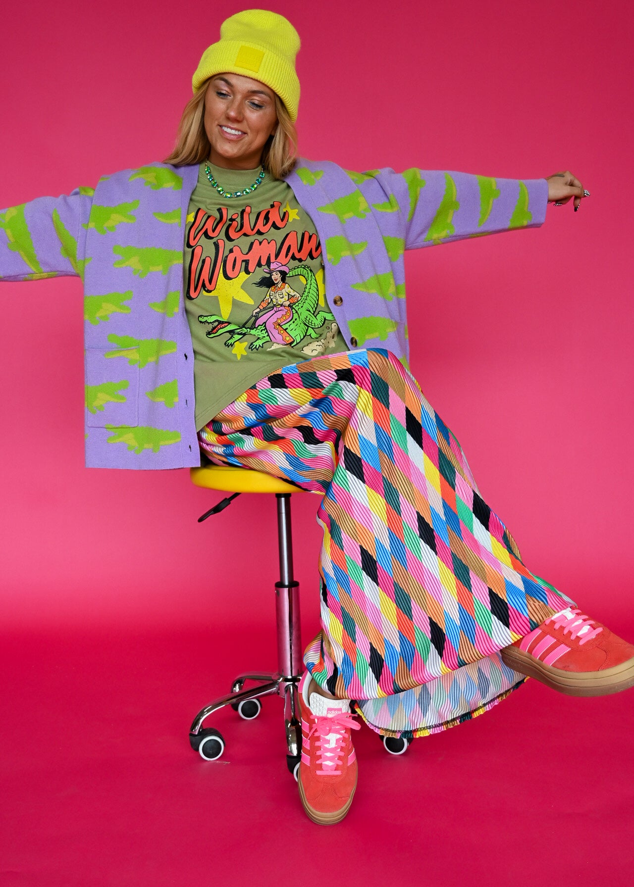 A woman posing on a yellow stool against a vibrant pink background, wearing a colorful retro-themed outfit with a purple cardigan, green graphic t-shirt, multicolor geometric pants, and a yellow beanie
