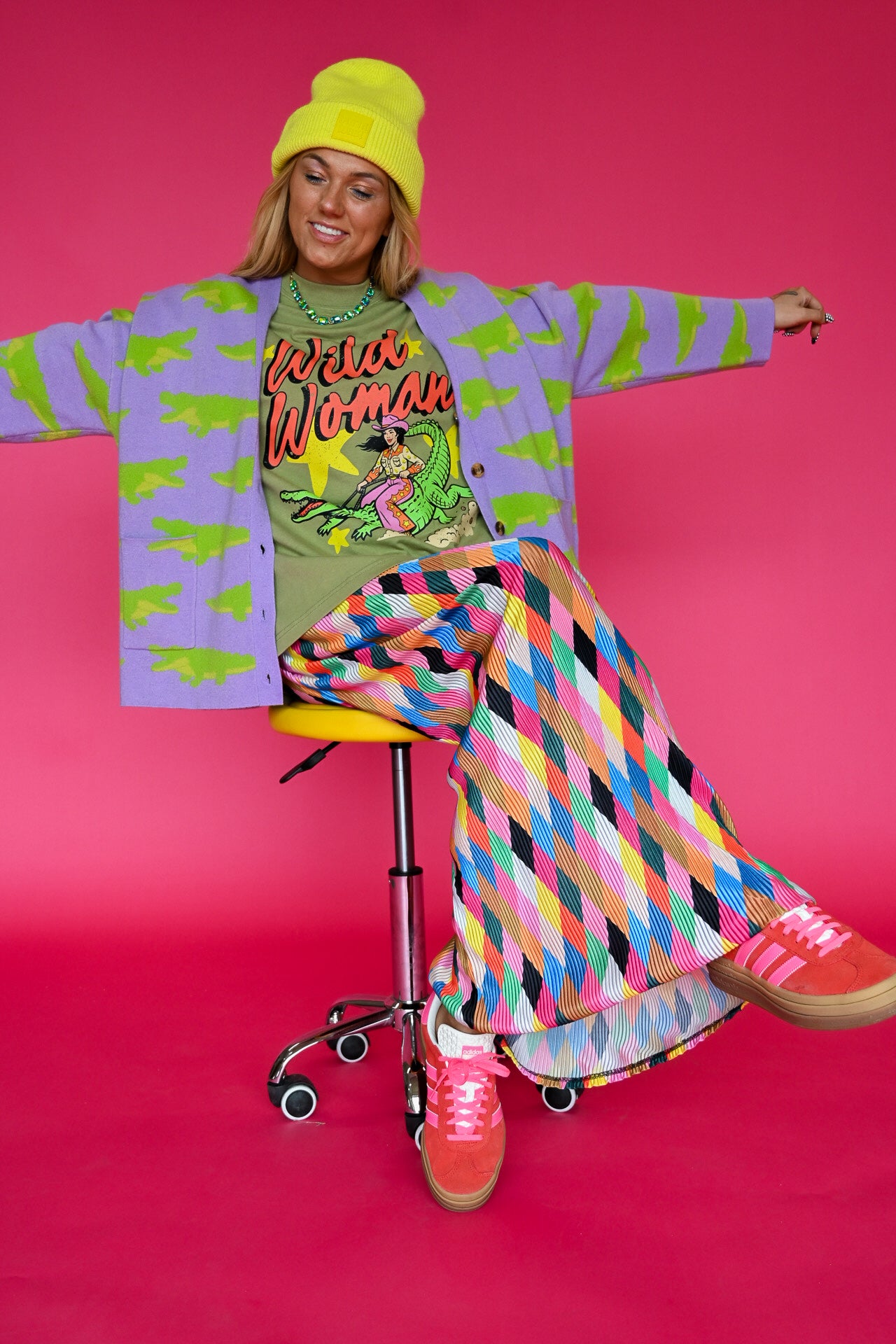A woman posing on a yellow stool against a vibrant pink background, wearing a colorful retro-themed outfit with a purple cardigan, green graphic t-shirt, multicolor geometric pants, and a yellow beanie