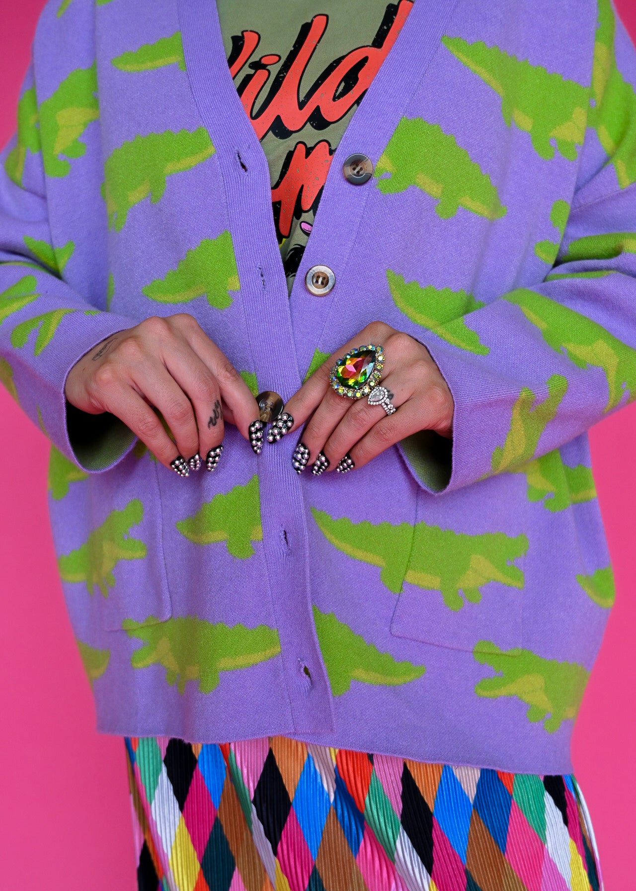 A close-up of a person wearing a lavender cardigan with a green crocodile pattern, layered over a colorful graphic shirt and a multicolored diamond-patterned skirt, set against a solid pink background