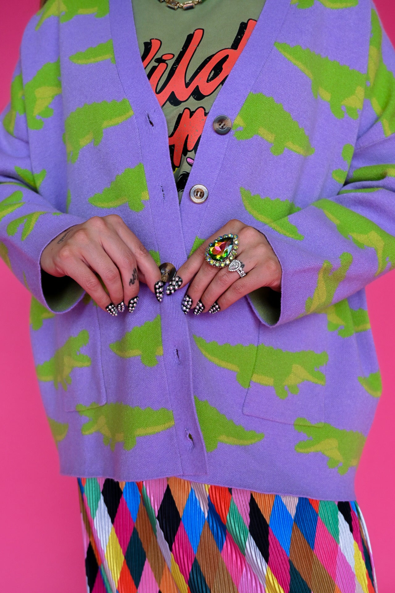 A close-up of a person wearing a lavender cardigan with a green crocodile pattern, layered over a colorful graphic shirt and a multicolored diamond-patterned skirt, set against a solid pink background