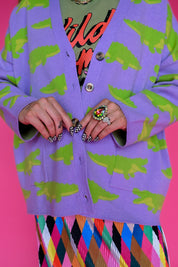 A close-up of a person wearing a lavender cardigan with a green crocodile pattern, layered over a colorful graphic shirt and a multicolored diamond-patterned skirt, set against a solid pink background