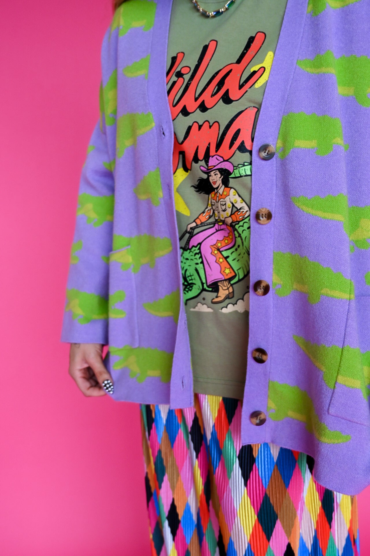 A person wearing a lavender cardigan with a green alligator print over a graphic t-shirt and a colorful argyle skirt, posed against a solid pink background
