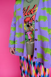 A person wearing a lavender cardigan with a green alligator print over a graphic t-shirt and a colorful argyle skirt, posed against a solid pink background