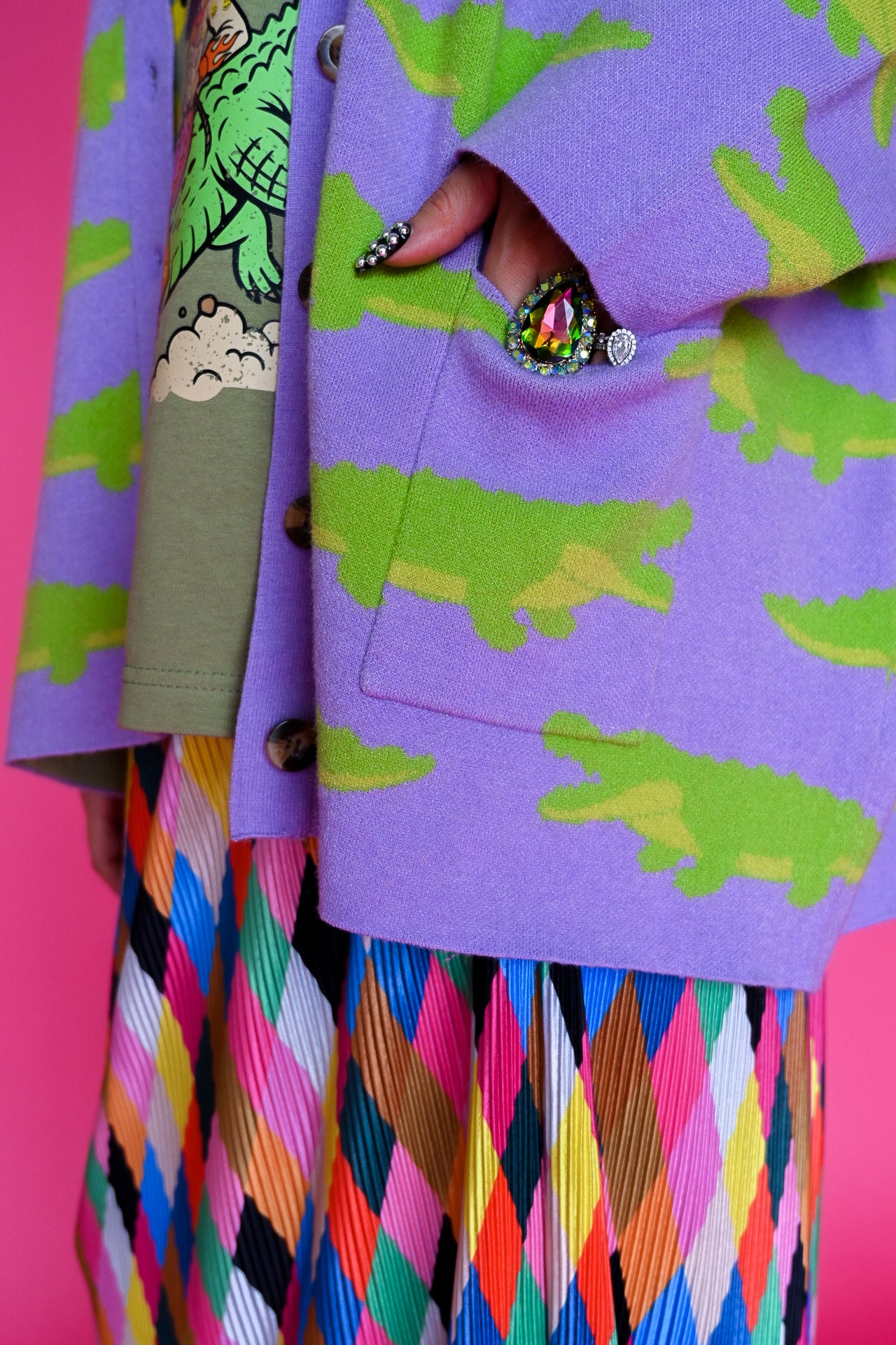 A close-up of a person wearing a lavender-purple cardigan with a green alligator print, layered over a colorful geometric-patterned pleated skirt. The background is a solid bright pink
