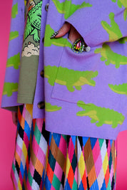 A close-up of a person wearing a lavender-purple cardigan with a green alligator print, layered over a colorful geometric-patterned pleated skirt. The background is a solid bright pink