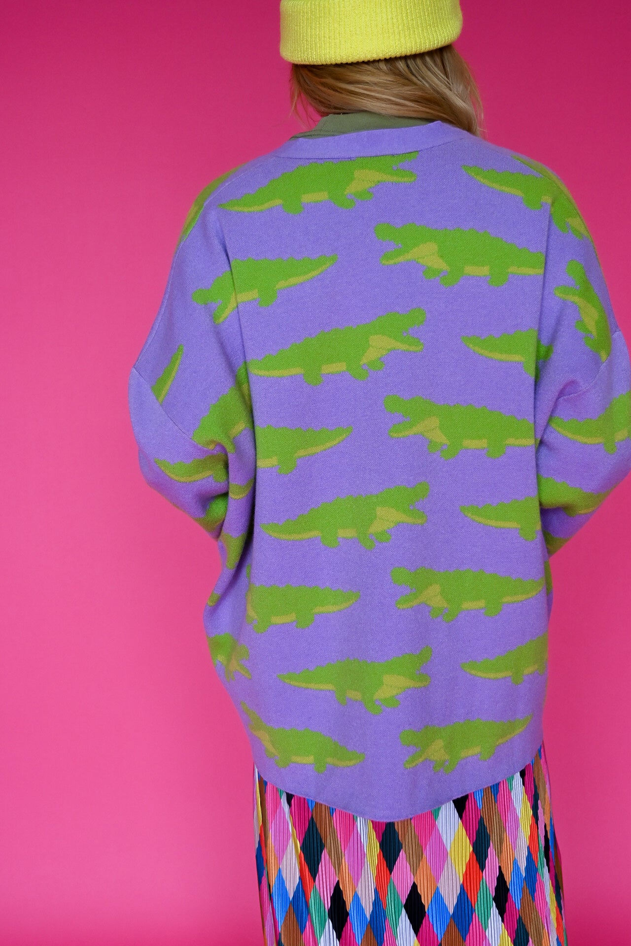 A person shown from the back wearing a purple knit cardigan with a green alligator print, paired with a yellow beanie and a colorful diamond-patterned skirt, set against a solid pink background