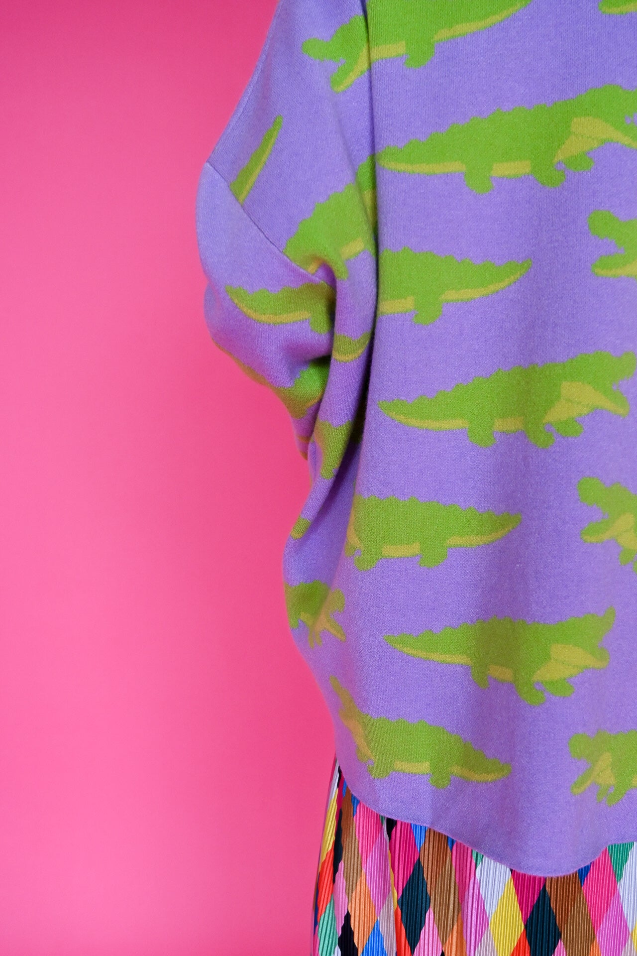 A close-up of a purple sweatshirt featuring a repeating pattern of bright green alligators, paired with a multicolored argyle skirt, set against a solid pink background