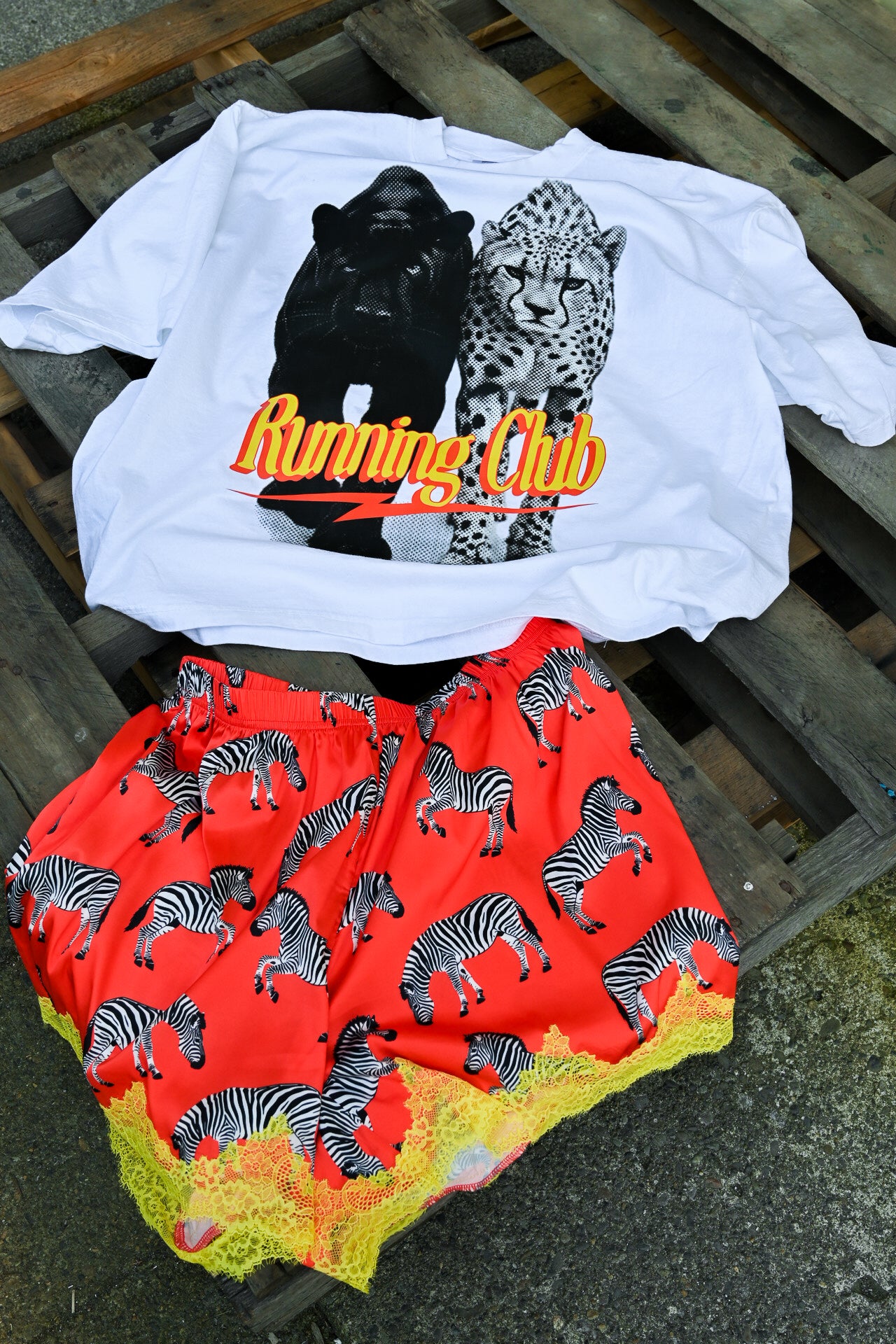 A white graphic T-shirt with a black panther and leopard print and the words 'Running Club' in orange, laid flat on a wooden pallet next to red shorts with a zebra pattern and yellow lace trim