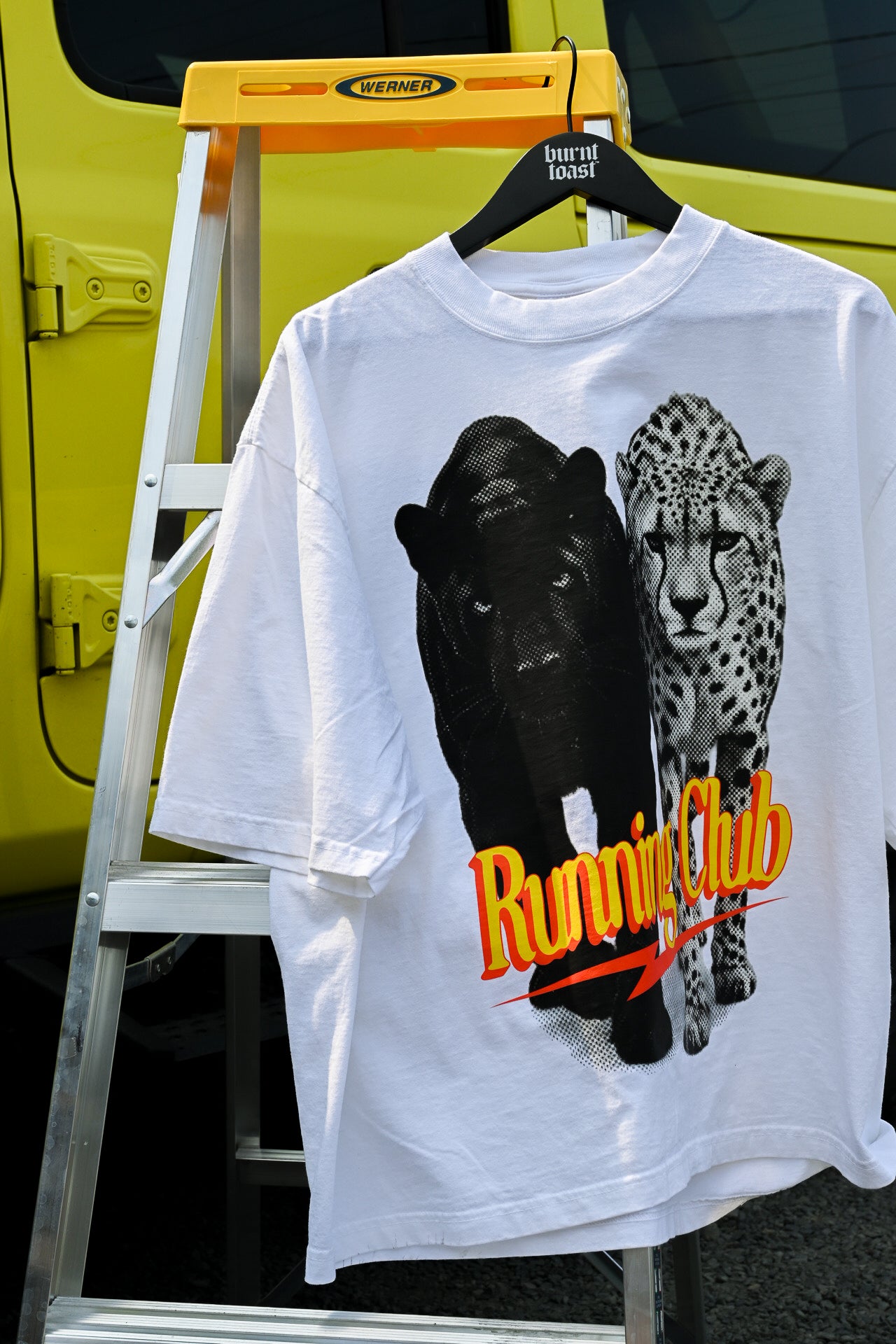 A white graphic T-shirt hanging on a black hanger from a yellow step ladder. The shirt features a large print of a black panther and a cheetah, with the text 'Running Club' in orange and yellow