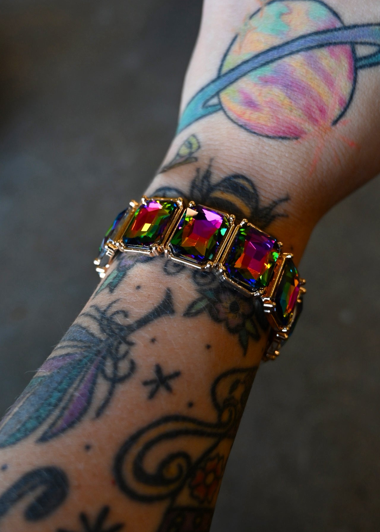 A multi-colored jewel bracelet on a models wrist with tattoos on a dark background