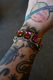 A multi-colored jewel bracelet on a models wrist with tattoos on a dark background