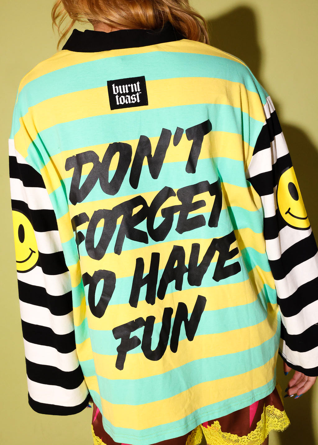 A person shown from the back wearing a burnt toast brand long-sleeve shirt with yellow and mint green horizontal stripes, black text reading 'DON'T FORGET TO HAVE FUN', black-and-white striped sleeves with yellow smiley faces, and a black collar.