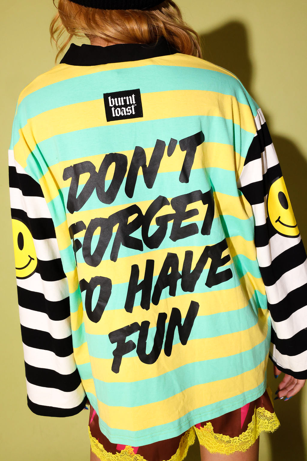 A person shown from the back wearing a burnt toast brand long-sleeve shirt with yellow and mint green horizontal stripes, black text reading 'DON'T FORGET TO HAVE FUN', black-and-white striped sleeves with yellow smiley faces, and a black collar.