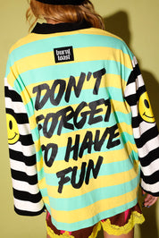 A person shown from the back wearing a burnt toast brand long-sleeve shirt with yellow and mint green horizontal stripes, black text reading 'DON'T FORGET TO HAVE FUN', black-and-white striped sleeves with yellow smiley faces, and a black collar.